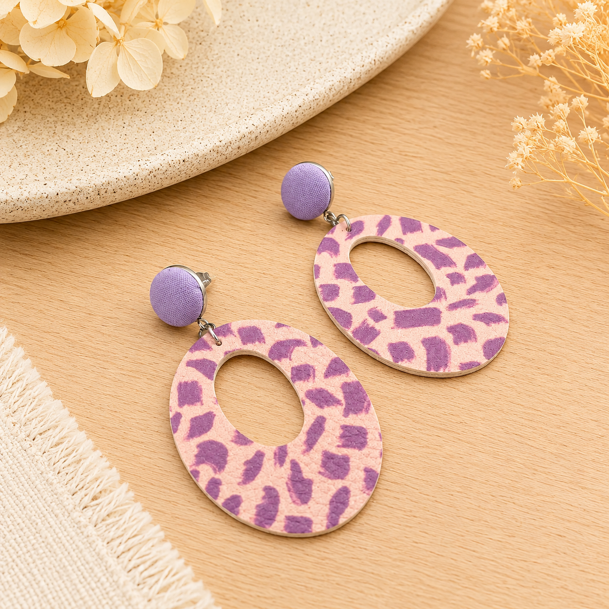 Handmade lilac and blush faux leather statement dangle earrings with abstract pattern and oval cut-out design styled on a light oak surface with soft neutral décor.