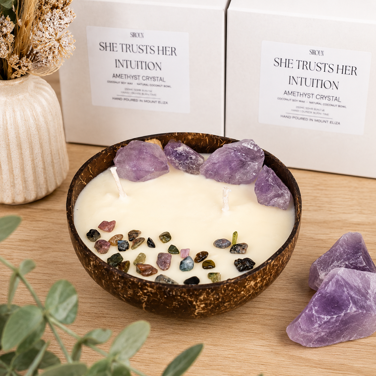 Handmade coconut soy wax candle featuring genuine amethyst crystals and gemstone chips in a natural coconut bowl, displayed on a light oak surface.