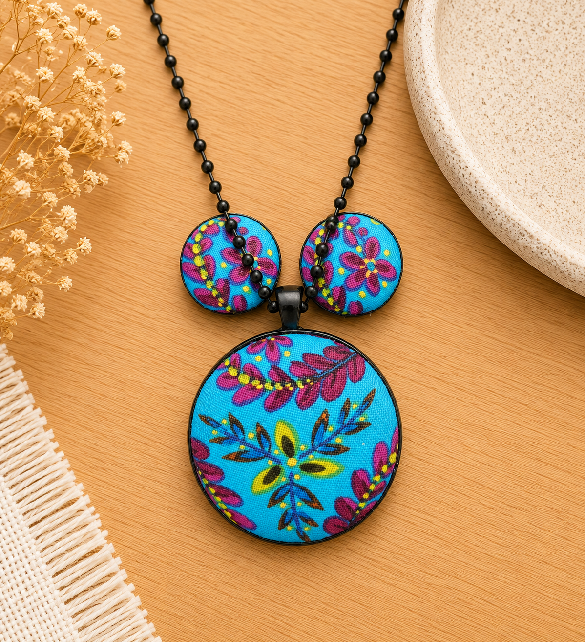 Handmade blue floral statement necklace and matching stud earrings displayed on a light oak surface in a soft neutral setting.