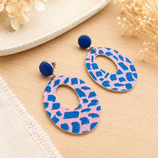 Handmade blue and pink faux leather statement dangle earrings with abstract pattern and oval cut-out design styled on a light oak surface with soft neutral décor.