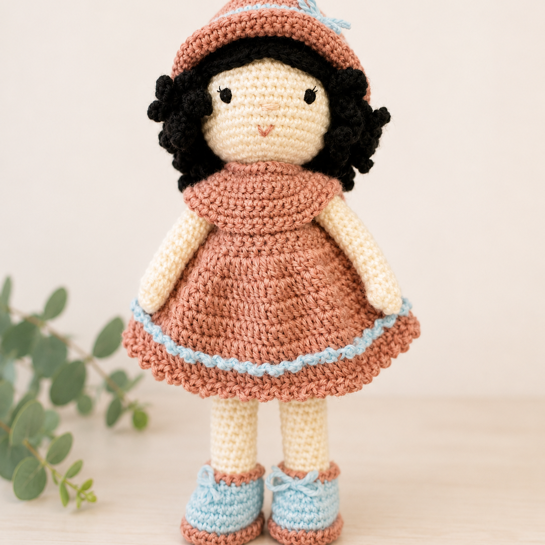 Handmade crochet doll wearing a blush pink dress with pastel blue trim and matching blue shoes.