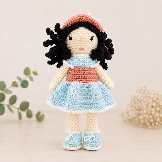 Handmade crochet doll with curly black hair wearing a pastel blue dress with blush pink details and matching blue shoes.