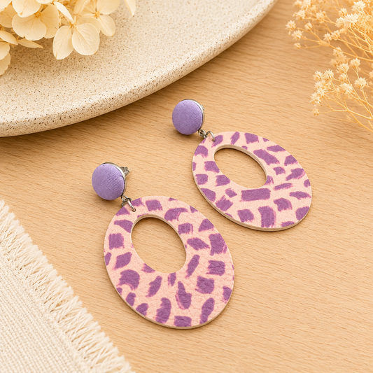 Handmade lilac and blush faux leather statement dangle earrings with abstract pattern and oval cut-out design styled on a light oak surface with soft neutral décor.