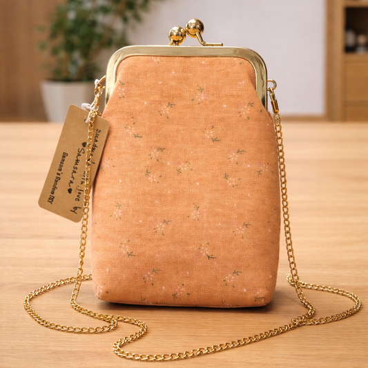 Peach mini clutch bag with subtle floral print and gold frame displayed on light oak background