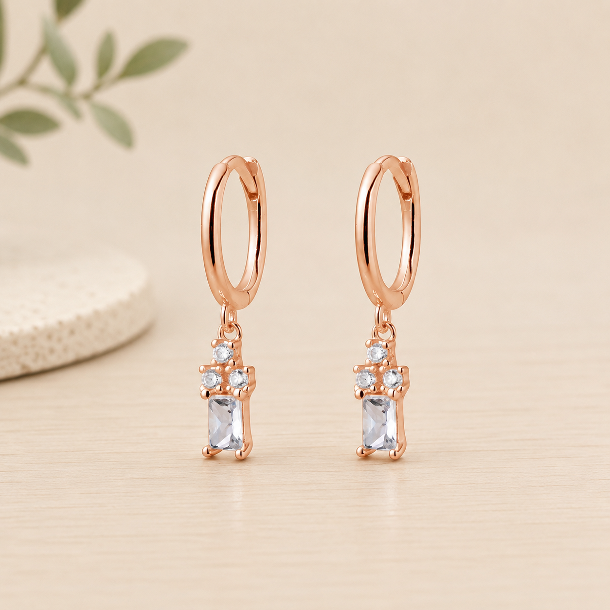 Rose gold sterling silver huggie earrings featuring a cubic zirconia trio with baguette drop charm on a neutral beige background.