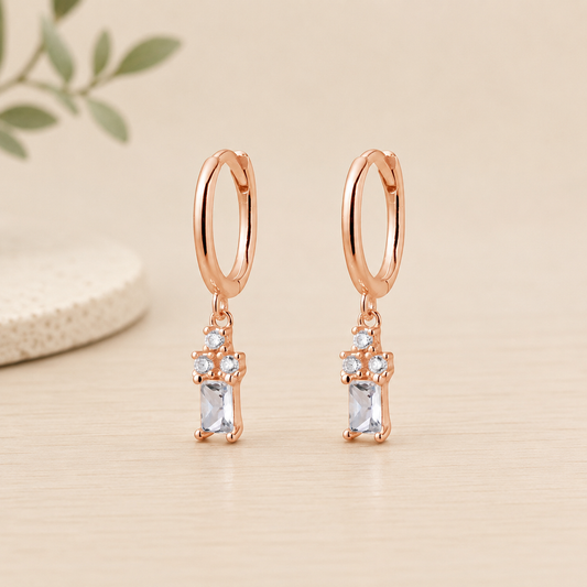 Rose gold sterling silver huggie earrings featuring a cubic zirconia trio with baguette drop charm on a neutral beige background.