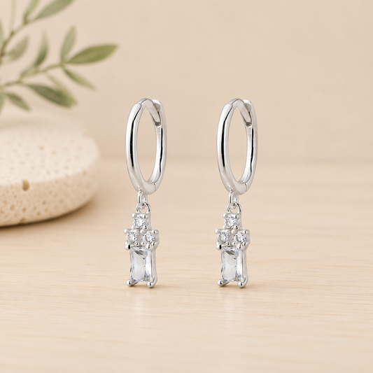Sterling silver huggie earrings featuring a cubic zirconia trio with baguette drop charm displayed on a neutral beige background.